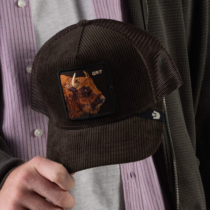 A person in a pink shirt and dark jacket holds the Corduroy Grit cap by Goorin Bros.—a dark brown corduroy hat featuring a bison patch with the word “GRIT,” finished with a curved brim and breathable mesh for enhanced comfort.