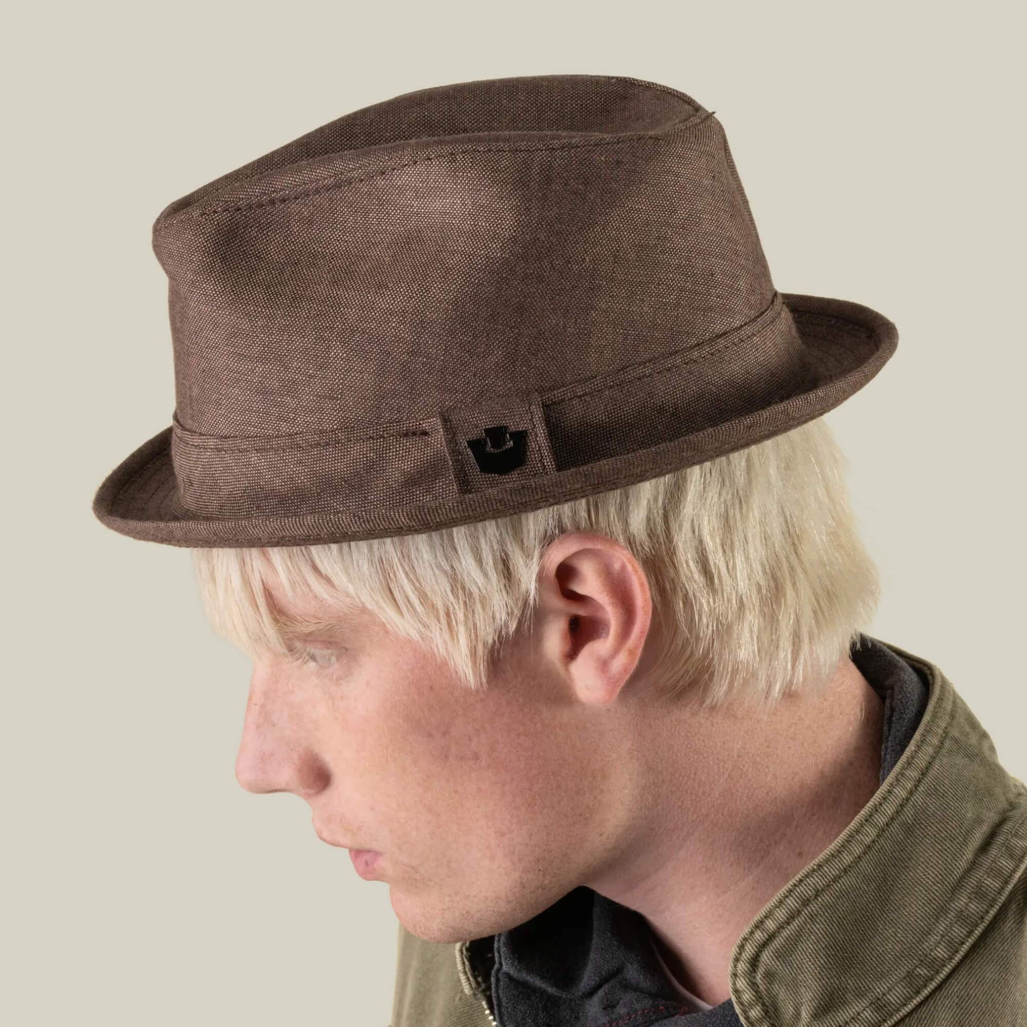 A person with light blond hair, wearing the Charlie Pants by Goorin Bros., a brown fedora hat, and an olive-green jacket, faces left against a plain beige background.