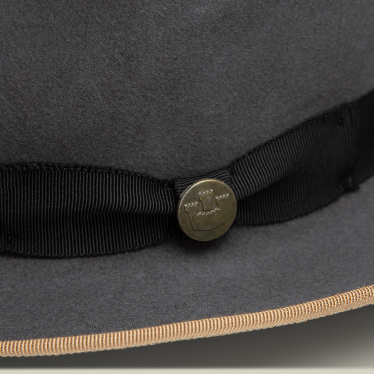 Close-up of the Goorin Bros. River Gray fedora, American made from merino wool felt in gray, featuring a black ribbon with a brass button etched design and a beige-trimmed wide brim.