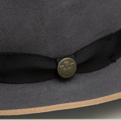 Close-up of the Goorin Bros. River Gray fedora, American made from merino wool felt in gray, featuring a black ribbon with a brass button etched design and a beige-trimmed wide brim.