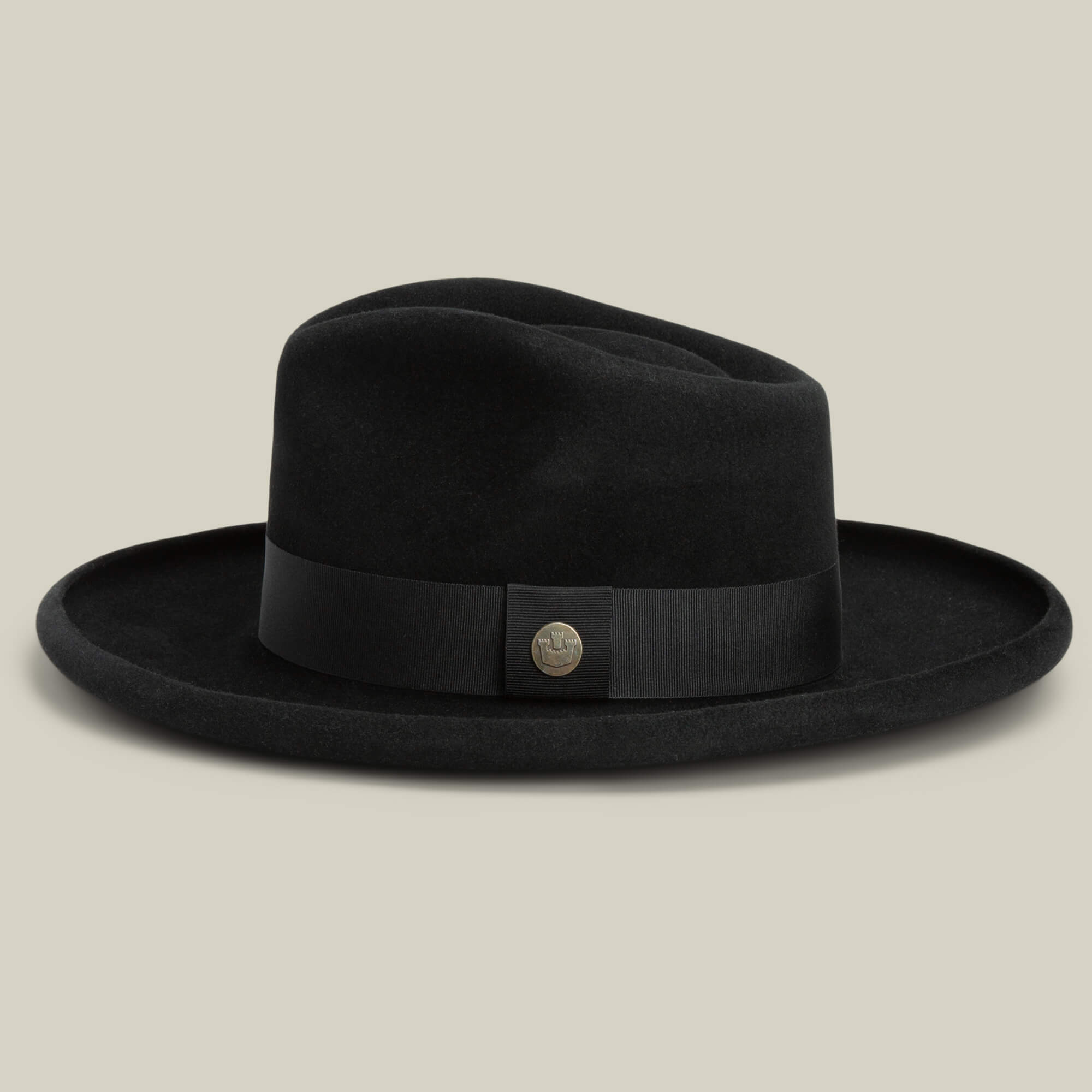 The Colonel Pierce by Goorin Bros. is a black wide-brim fedora with a teardrop crown, black ribbon band, and round metallic badge, displayed against a plain beige background.