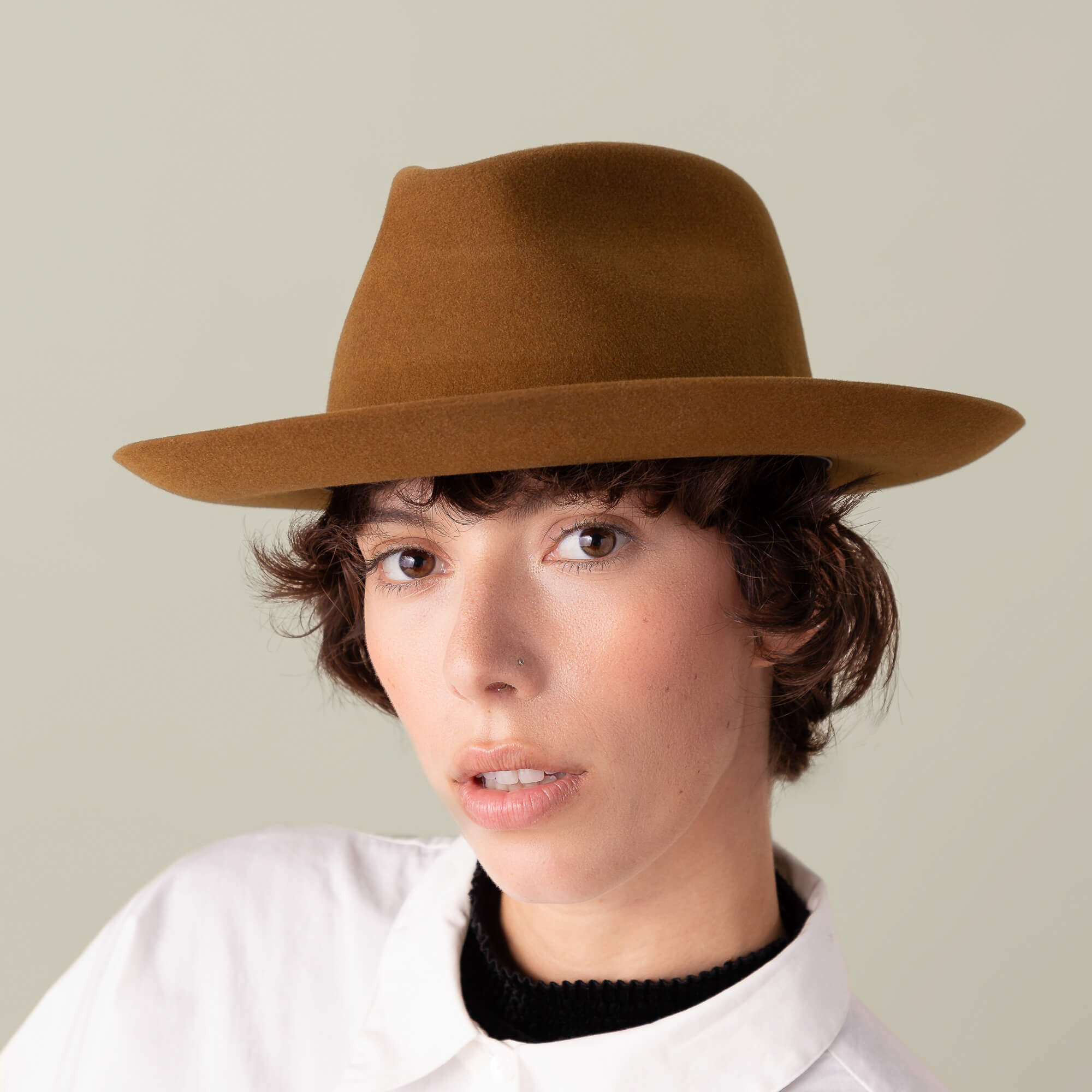 A person with short, curly brown hair wears the Goorin Bros. Night Hawk brown teardrop crown fedora with a wide brim and a white collared shirt, set against a neutral background.