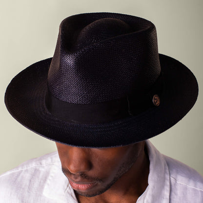 A man wears the Goorin Bros. John Sr., a black toquilla fedora hat with 50 UPF sun protection, and a white shirt, looking downward against a neutral background.