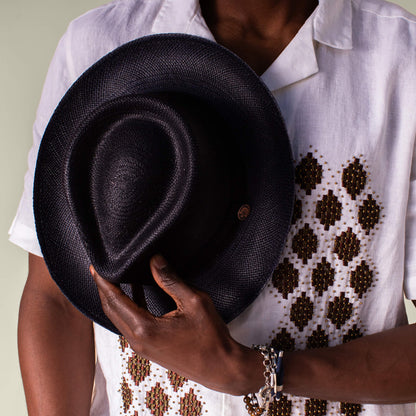 A person in a white patterned shirt holds the Goorin Bros. John Sr. black toquilla fedora (50 UPF) in front of their chest.