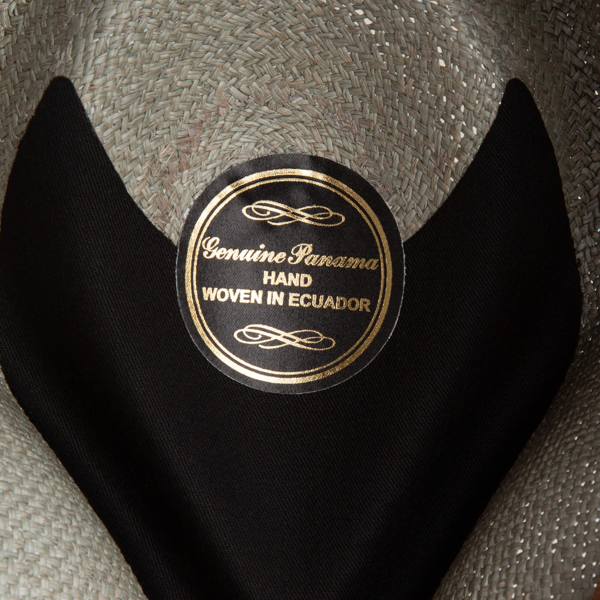 Close-up of a hat label reading Genuine Panama Hand Woven in Ecuador in gold lettering on a black background, showcasing the craftsmanship and 50 UPF sun protection of the John Sr. toquilla teardrop fedora by Goorin Bros.
