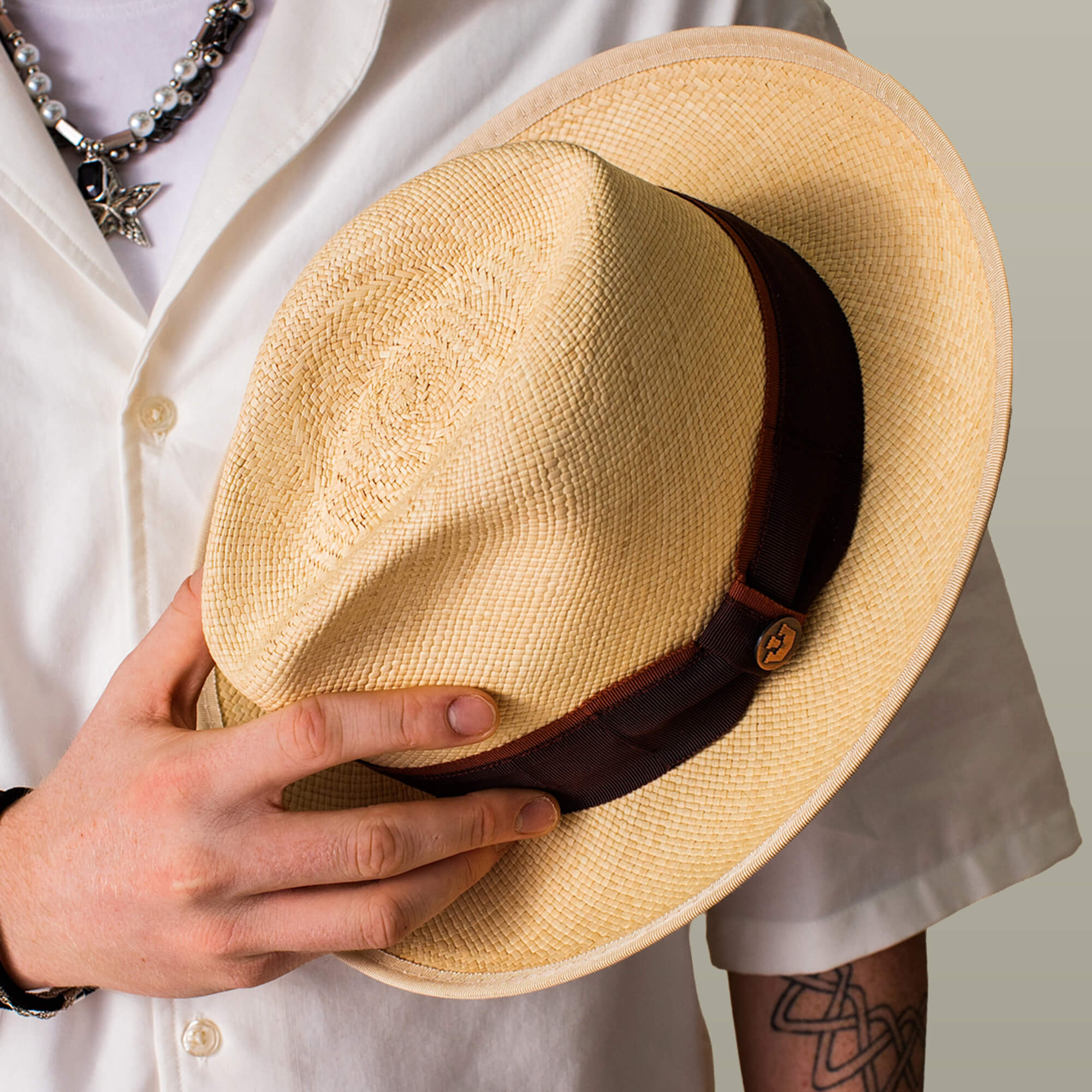 A person wearing a white shirt and jewelry holds the Goorin Bros. Hartman beige straw fedora with a dark brown grosgrain band.