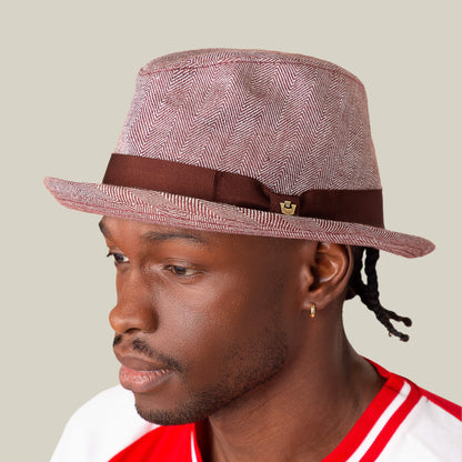 A man wearing a red and white shirt and the Zig Zag Big Stepper fedora by Goorin Bros., featuring a brown band and subtle herringbone pattern, faces left against a plain background.