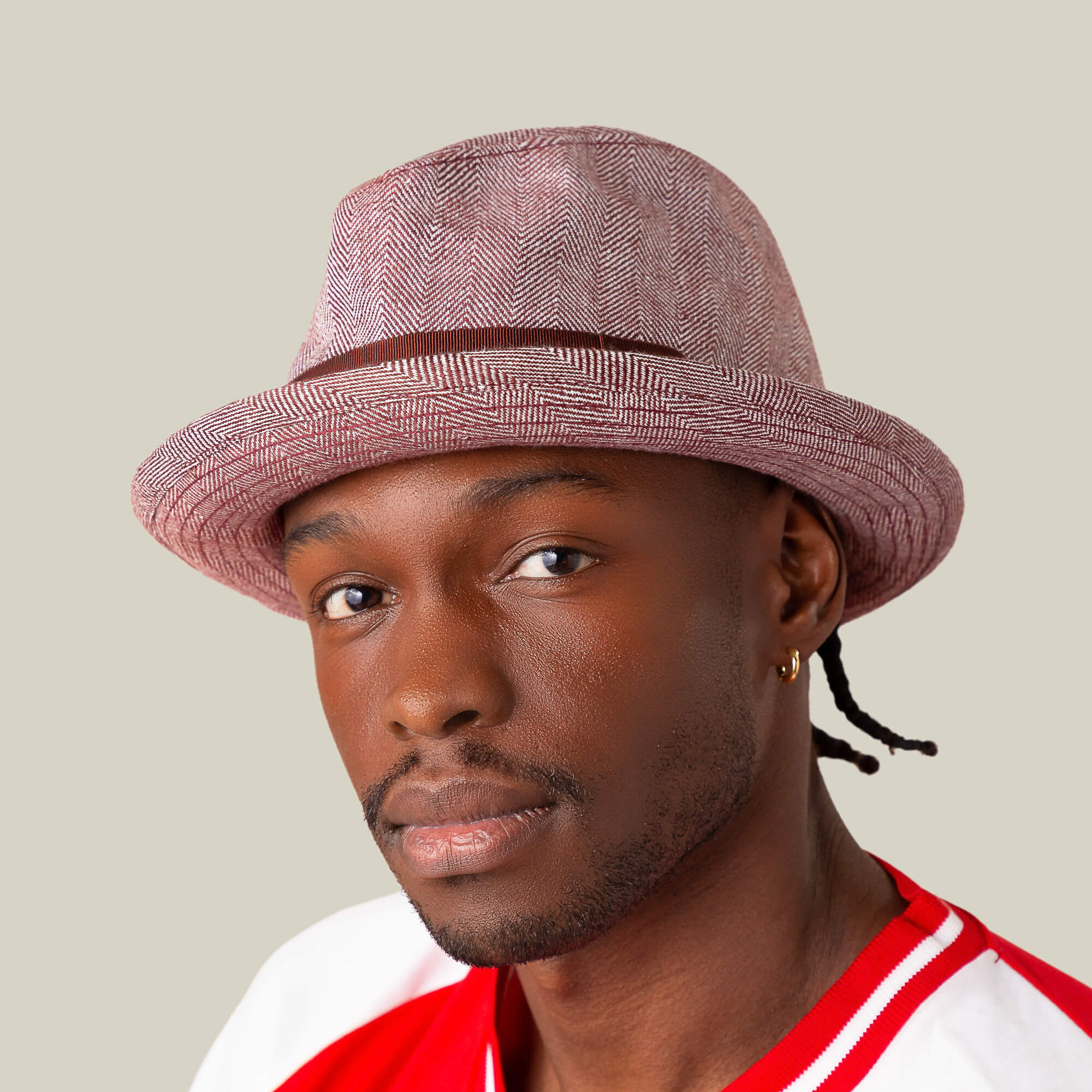 A man wearing the Goorin Bros. Zig Zag Big Stepper fedora with a light red herringbone pattern, a red and white shirt, and a small hoop earring looks at the camera against a plain background.
