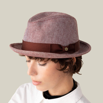 A person wears the Goorin Bros. Zig Zag Big Stepper—a pink structured fedora with a herringbone pattern, brown ribbon, and pin—looking down against a plain beige background.