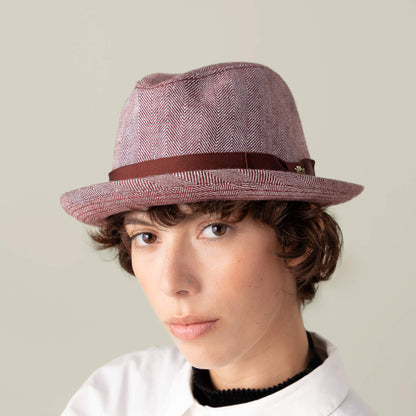 Person with short curly hair wears the Goorin Bros. Zig Zag Big Stepper, a light red structured fedora with a brown herringbone ribbon, paired with a white collared shirt and a neutral expression against a plain background.