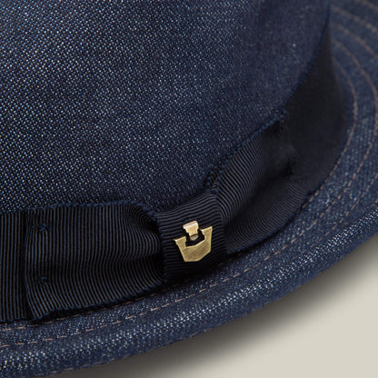 Close-up of the Goorin Bros. Noisy Big Stepper fedora in dark blue denim, highlighted by a sharp crown, black grosgrain ribbon, and a small gold metal emblem for a refined finish.