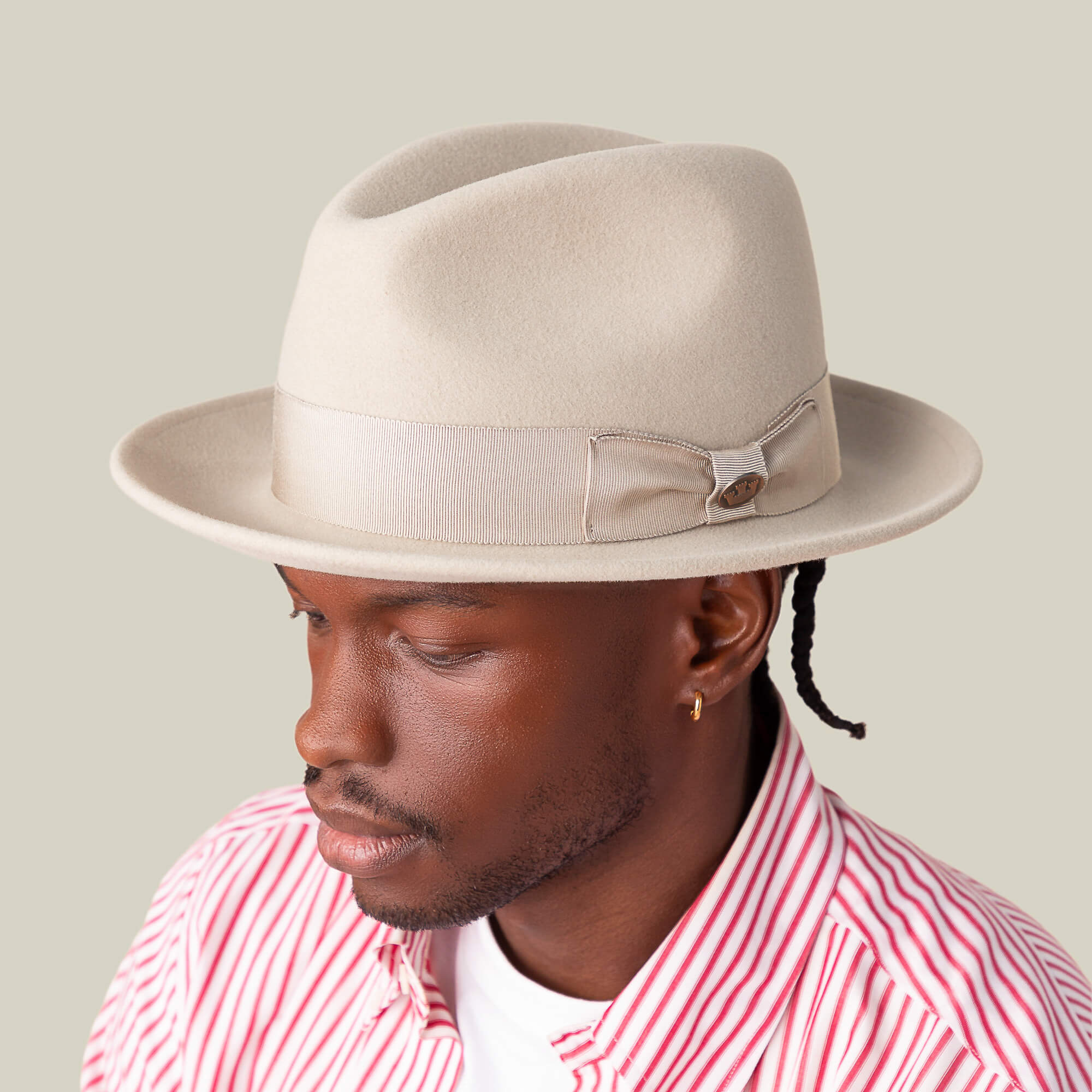 A man wears the Goorin Bros. "Dean the Butcher" fedora with a beige bow and German wicking sweatband, paired with a gold hoop earring and red-and-white striped shirt, looking downward against a neutral background.
