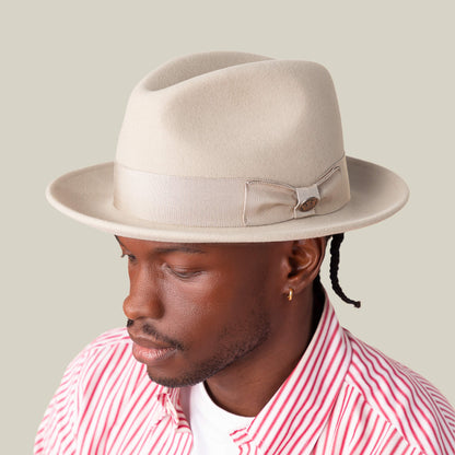 A man wears the Goorin Bros. "Dean the Butcher" fedora with a beige bow and German wicking sweatband, paired with a gold hoop earring and red-and-white striped shirt, looking downward against a neutral background.