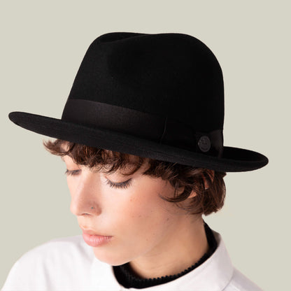 A person with short hair and a nose piercing wears the Dean the Butcher fedora by Goorin Bros., featuring a snap brim, paired with a white collared shirt, and looks down against a plain background.