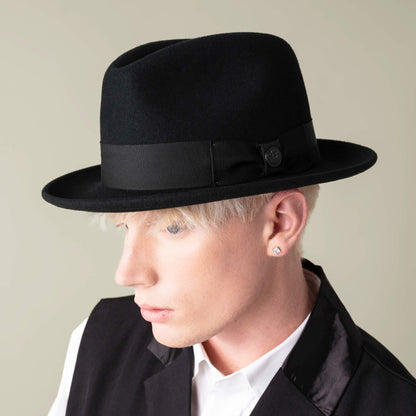 A blond-haired person wears the Dean the Butcher fedora by Goorin Bros., featuring a snap brim, paired with a black vest and white shirt, looking down and to the left against a neutral background.