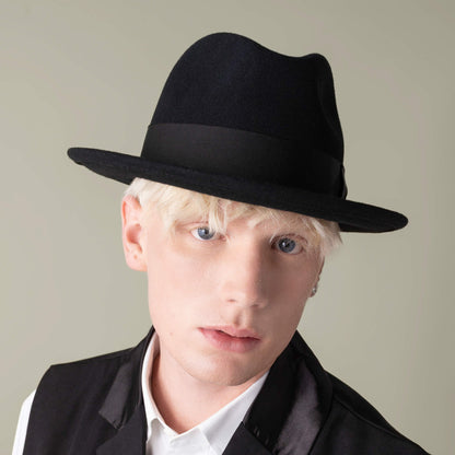 A young person with light blonde hair wears the "Dean the Butcher" fedora by Goorin Bros., styled with a black vest and white shirt, looking directly at the camera against a neutral background.