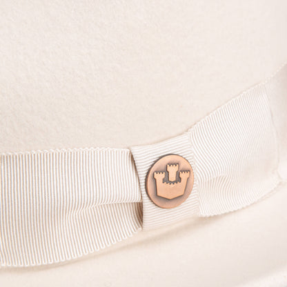 Close-up of the Goorin Bros. “Dean the Butcher” fedora in cream, featuring a matching grosgrain hatband and a round metal badge with castle emblem—an elegant hat for any season.