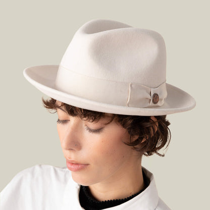 A person with short curly hair wears Goorin Bros.' "Dean the Butcher" fedora, featuring a grosgrain hatband and button detail, paired with a white collared shirt, looking downward against a plain background.