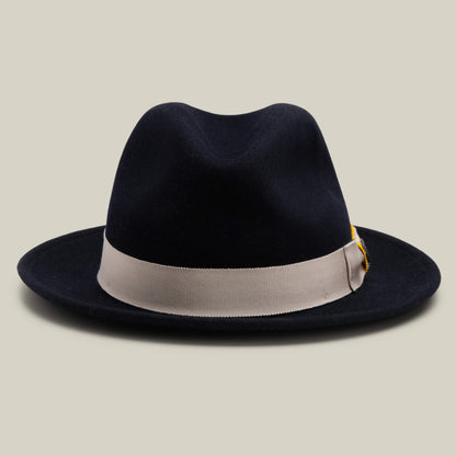 Dean the Butcher by Goorin Bros. is an EDGE-colored American-made fedora, featuring a wide light gray ribbon band, German wicking sweatband, and a small feather accent, displayed against a plain beige background.