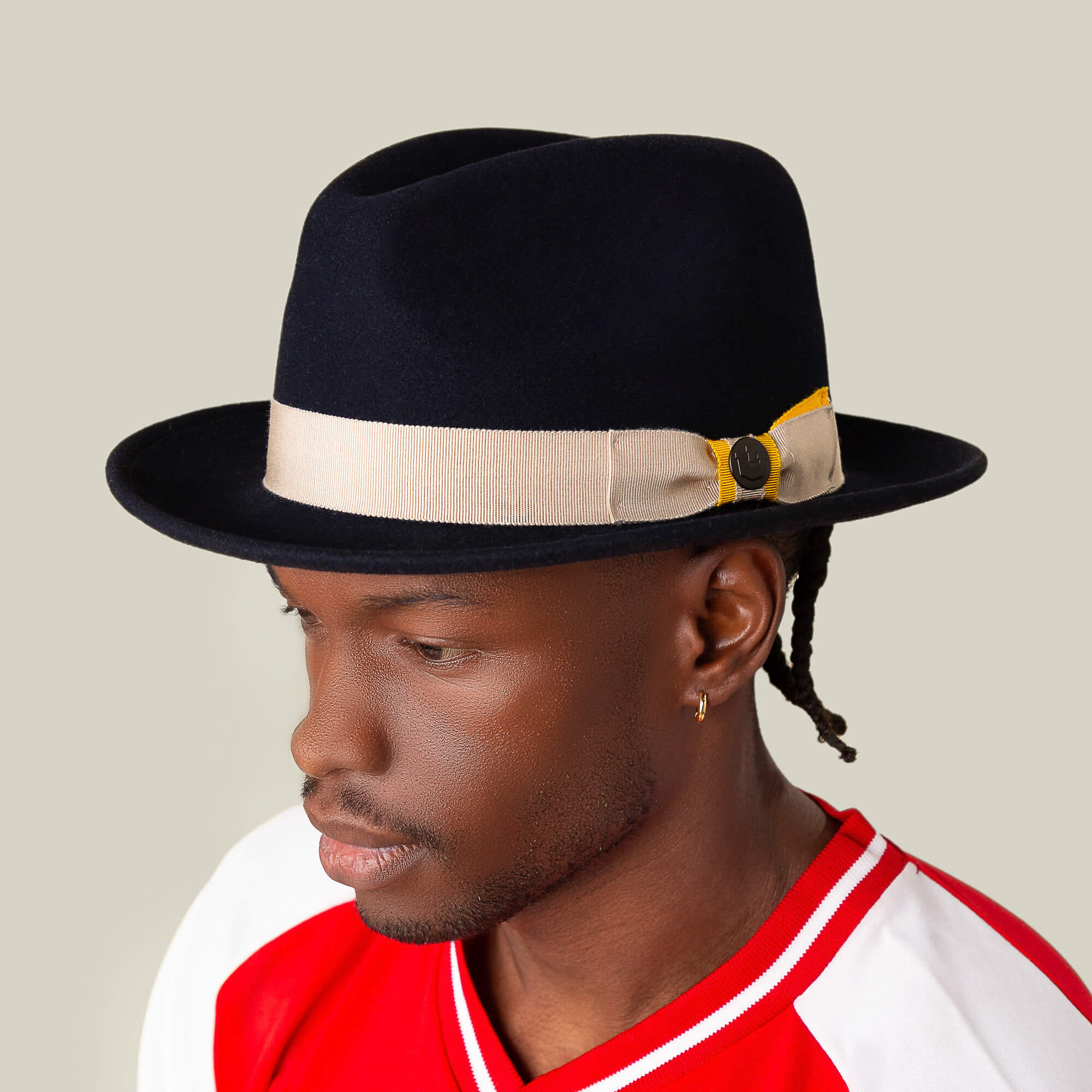 A man wearing the Goorin Bros. "Dean the Butcher"—a black, American-made fedora with a beige band and button—pairs it with a red-and-white shirt, facing slightly left against a plain background.