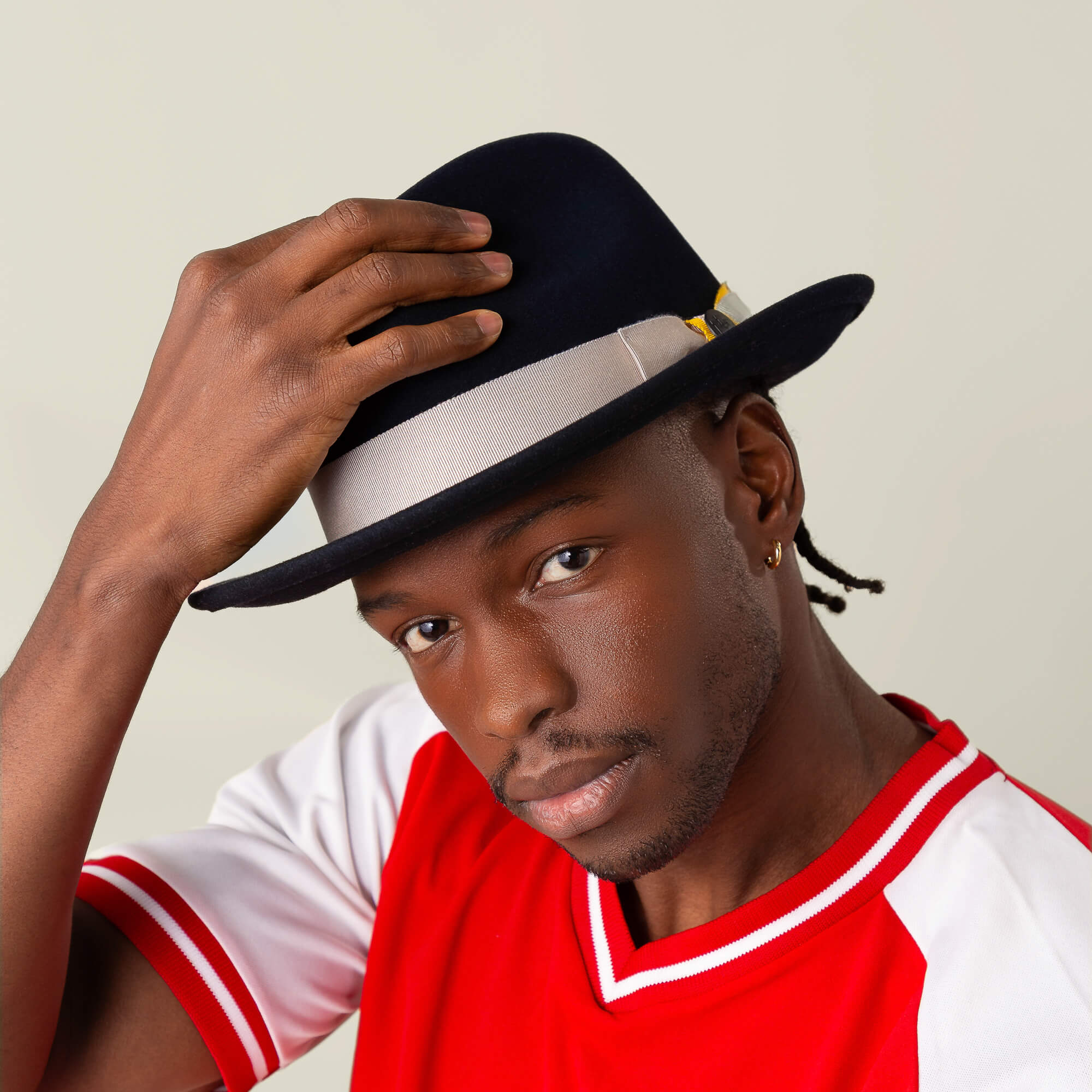 A man in a red and white shirt adjusts the Dean the Butcher fedora by Goorin Bros., featuring a gray band and German wicking sweatband, while looking toward the camera against a plain background.