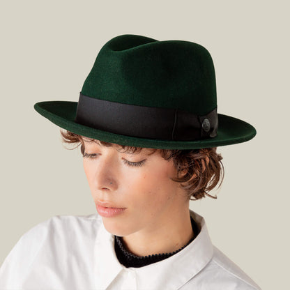 A person wearing the Goorin Bros. Dean the Butcher dark green fedora with a black band and a white collared shirt, looking downward against a plain light background.