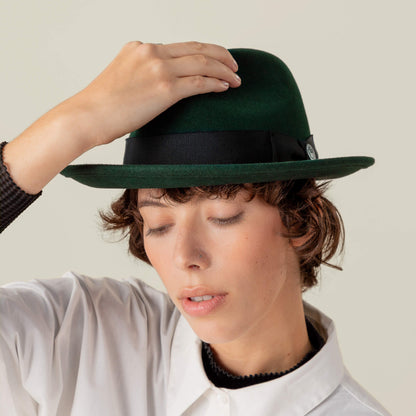 A person wears the Dean the Butcher dark green fedora by Goorin Bros. and a white collared shirt, eyes closed, with one hand touching the top of the hat.