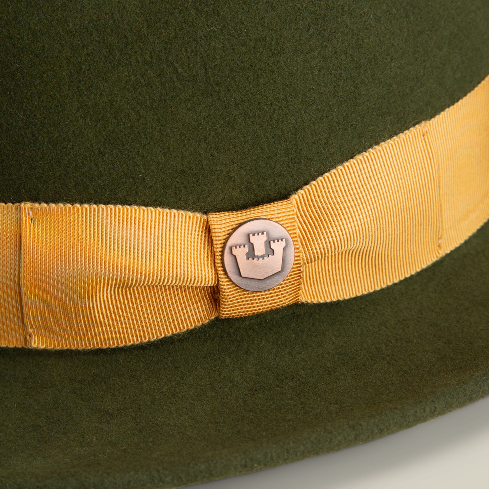 Close-up of Dean the Butcher by Goorin Bros., an all-season green felt hat featuring a yellow grosgrain band with a central castle emblem pin.