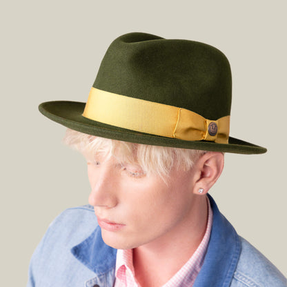A person with light blonde hair wears the "Dean the Butcher" olive green fedora by Goorin Bros., featuring a yellow band, paired with a light denim jacket, pink checked shirt, and a stud earring, set against a plain background.
