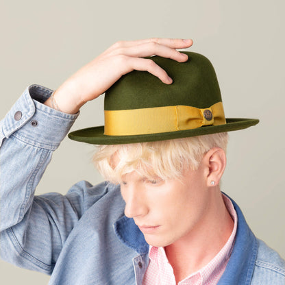A person with light blonde hair tilts the Goorin Bros. "Dean the Butcher" green fedora with a yellow band, paired with a denim jacket.