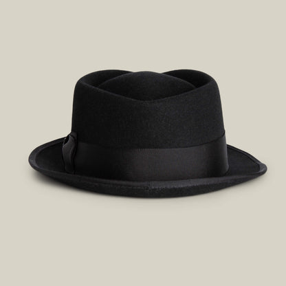 The Goorin Bros. Slick Carter is a black wool short brim pork pie hat with a wide grosgrain ribbon band, shown against a plain beige background, embodying effortless style.