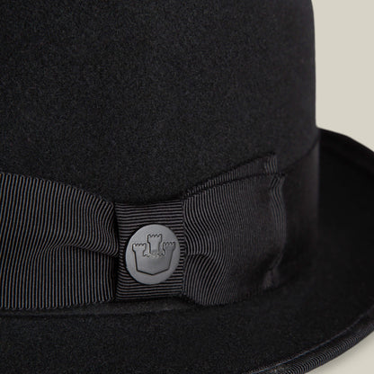 Close-up of the Slick Carter by Goorin Bros.: a black felt hat with a ribbon and round metal pin featuring a castle turret emblem, showcasing effortless style and a classic wool short brim design.