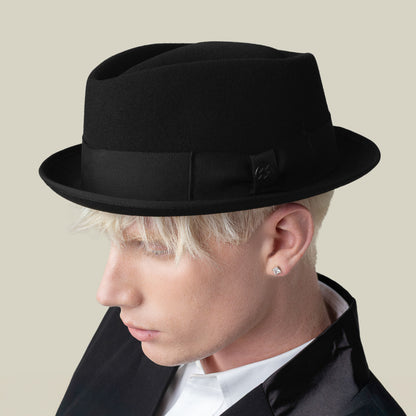 Wearing the Goorin Bros. Slick Carter black wool short brim fedora, a person with short blonde hair and a black blazer exudes effortless style in side profile against a plain background.
