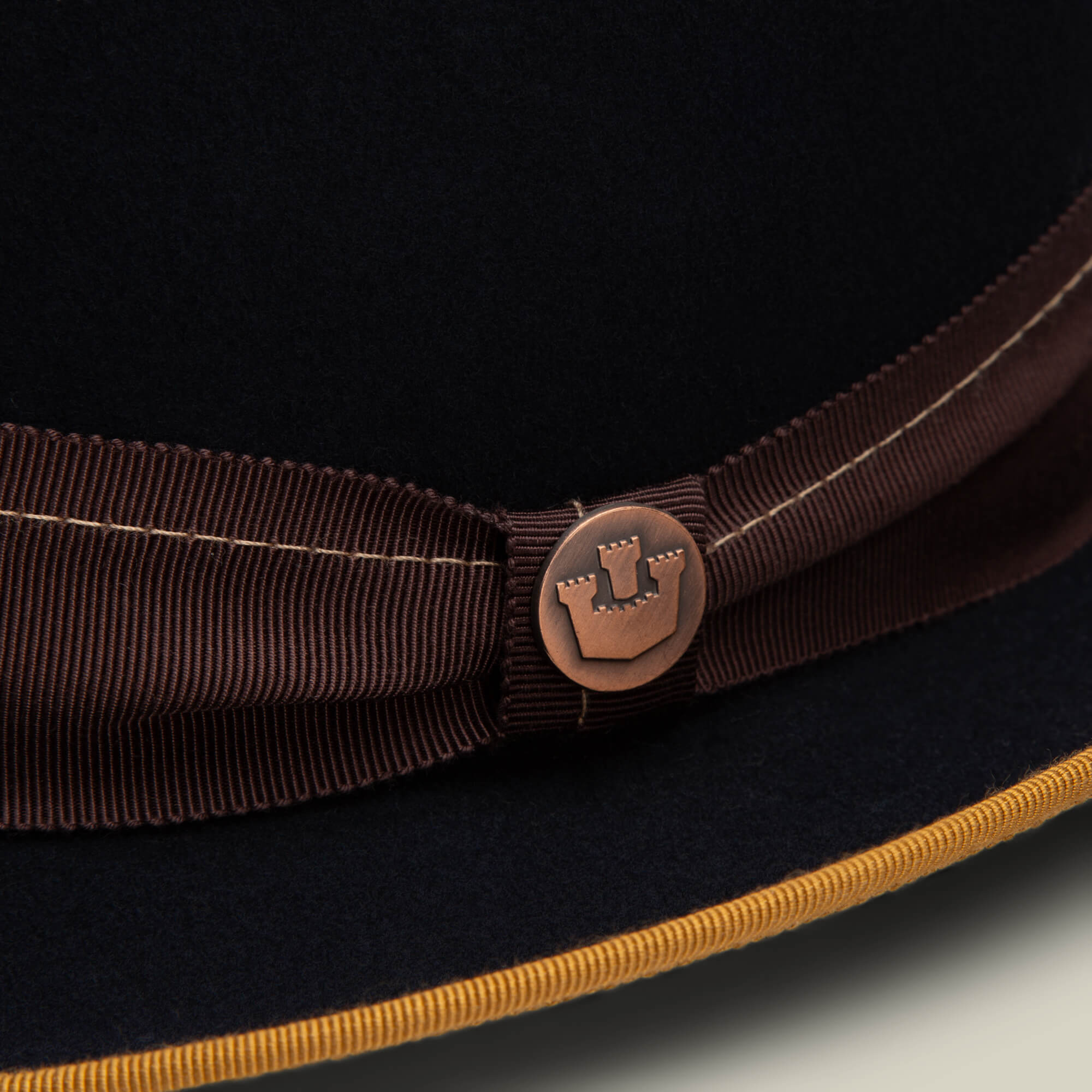 Close-up of the Slick Carter by Goorin Bros.: a wool short brim dark hat with a brown ribbon band, round metallic button embossed with a castle emblem, and yellow-trimmed edge for effortless style.