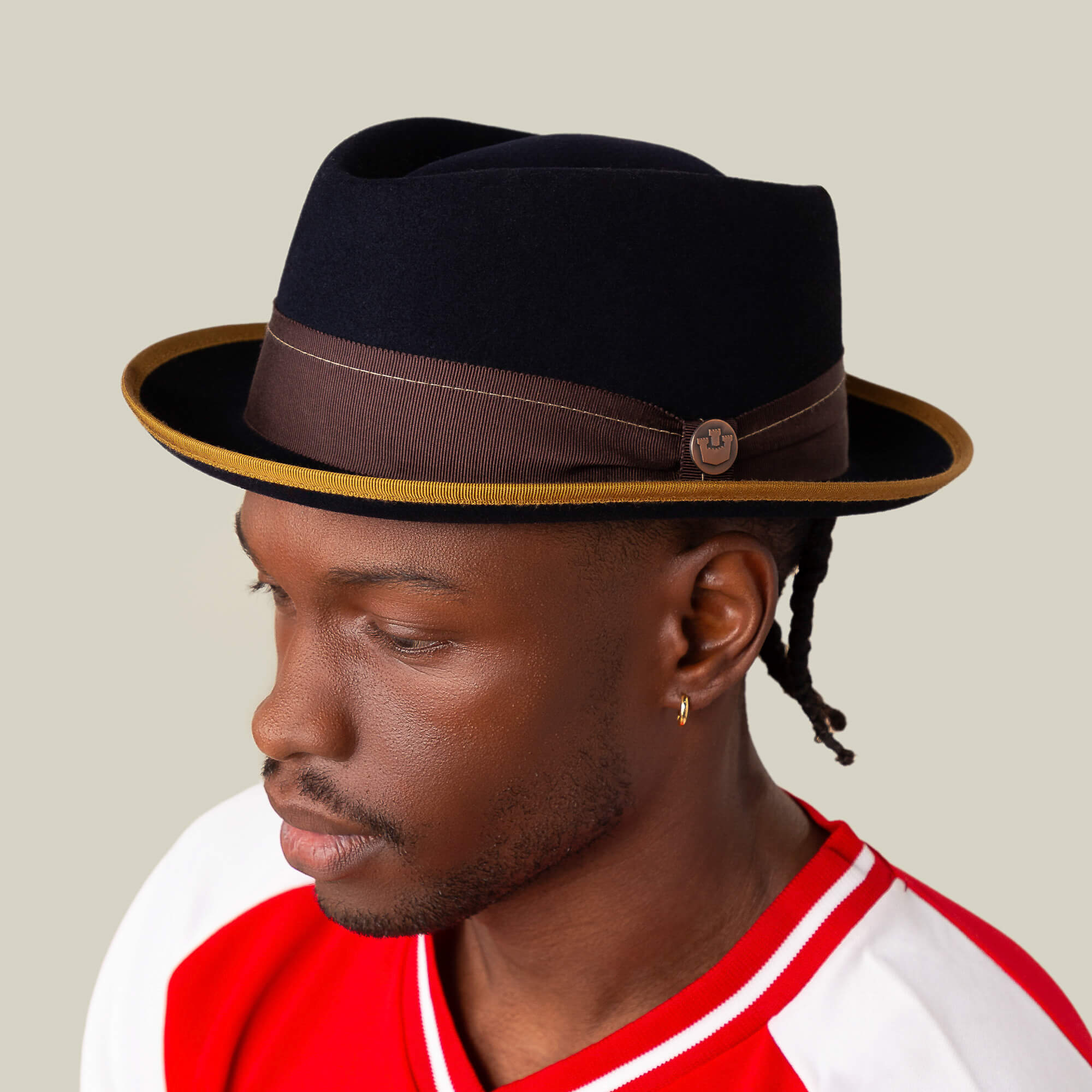 A man in a red and white shirt wears the Goorin Bros. Slick Carter short brim fedora, featuring a black wool finish with a brown band and gold trim, looking to the side against a plain background, exuding effortless style.