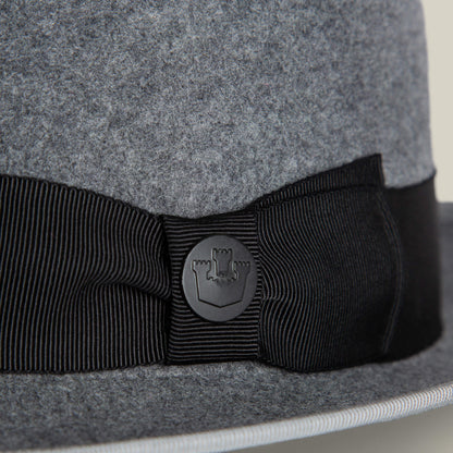 Close-up of the Slick Carter by Goorin Bros.: a grey wool short brim felt hat with a black ribbon and round badge featuring a shield and castle design, capturing effortless style.
