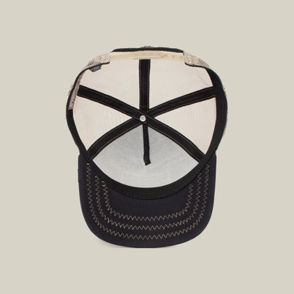 Inside view of The Cock by The Farm, a black and beige mesh trucker hat—its stitching and interior structure evoke bold character, echoing the rooster’s symbolism, all set against a plain background.