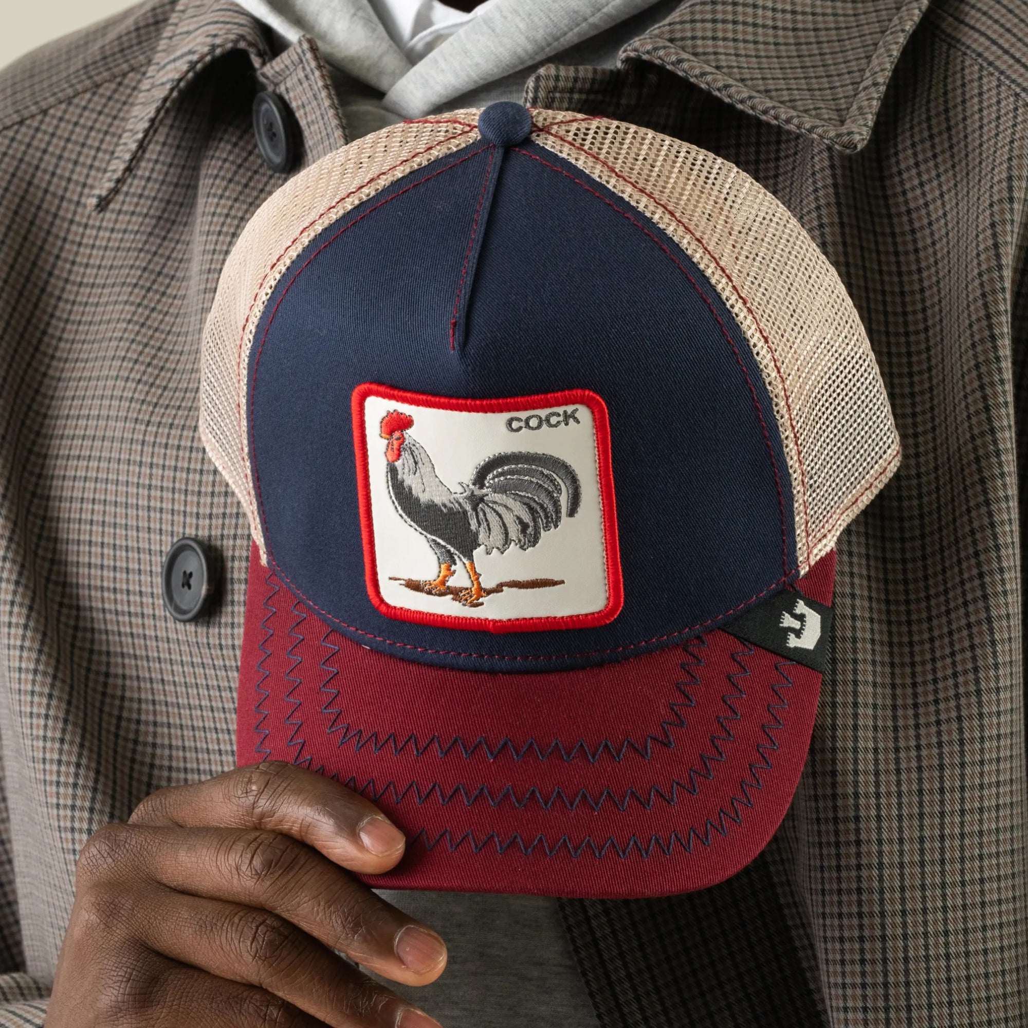A person holds The Farm’s “The Cock” baseball cap in navy and burgundy, featuring a rooster patch labeled COCK. They are dressed in a plaid jacket and gray hoodie.