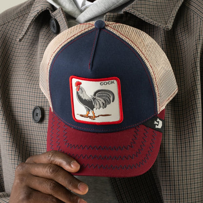 A person holds The Farm’s “The Cock” baseball cap in navy and burgundy, featuring a rooster patch labeled COCK. They are dressed in a plaid jacket and gray hoodie.