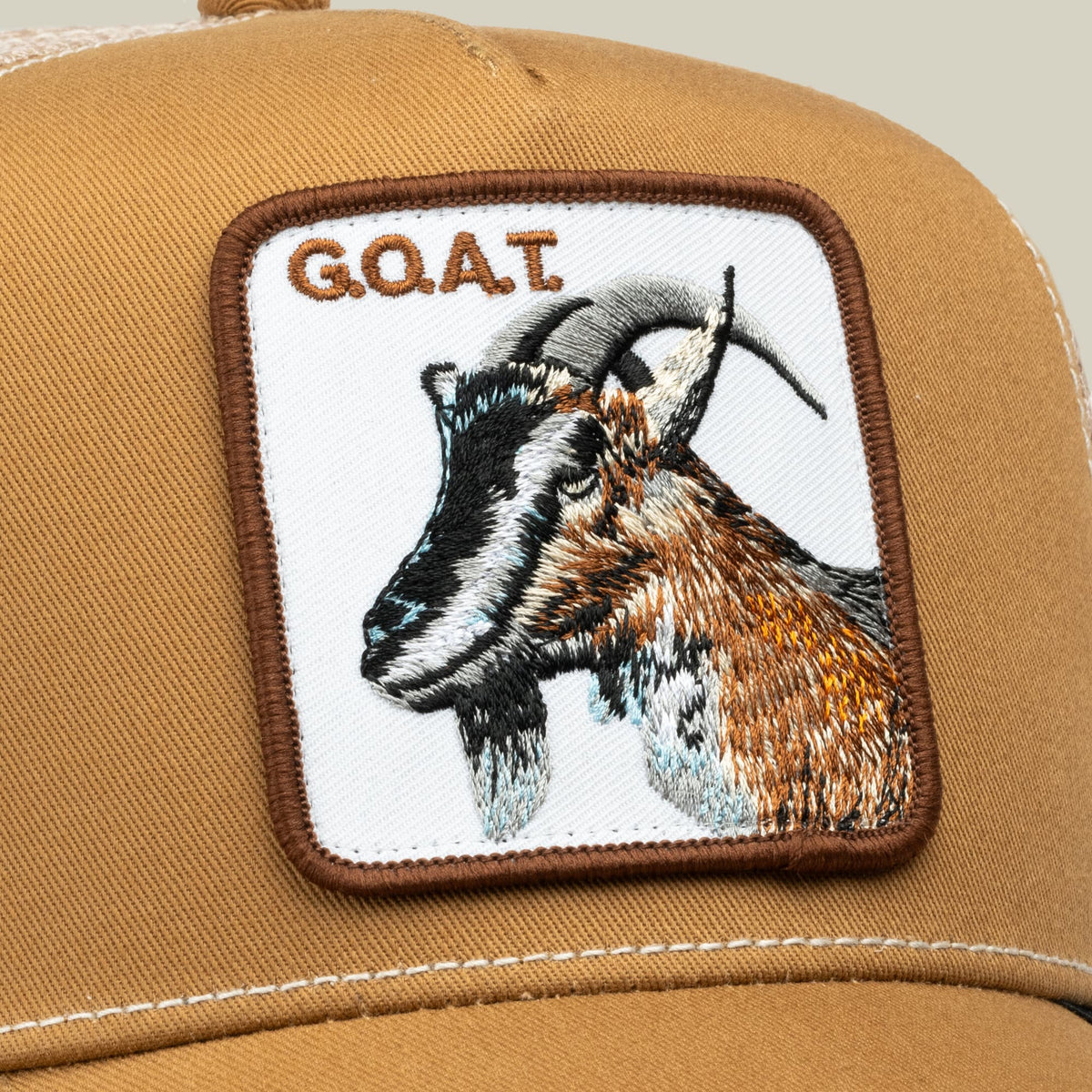 The GOAT by Goorin Bros is a tan trucker hat featuring an embroidered goat patch with brown and black detailing and the word GOAT in orange text on the front panel.