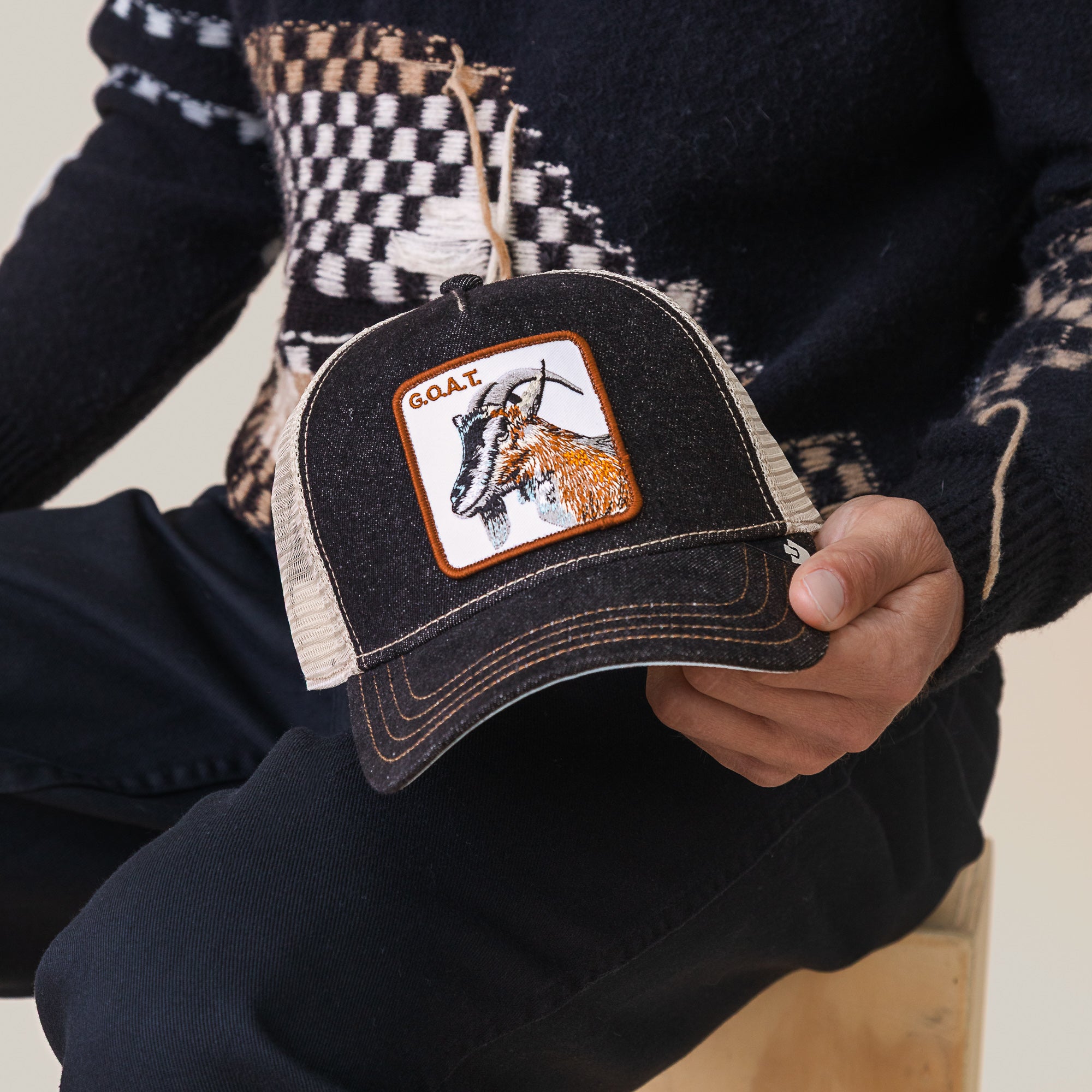 A person wears The Farm’s “The GOAT” hat—a black trucker with a goat patch and “G.O.A.T.” label—paired with a patterned black sweater and black pants.