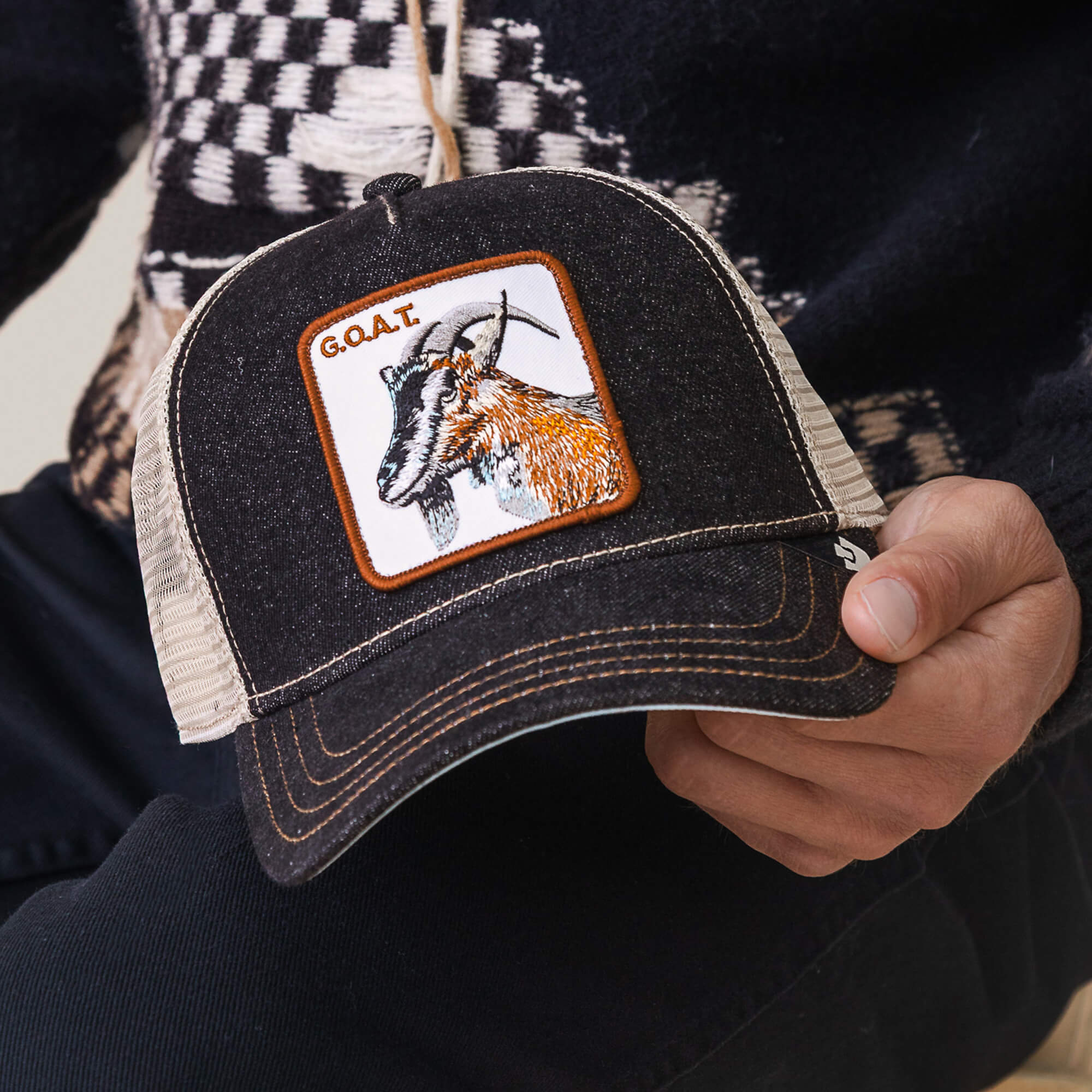 A person holds The Farm's black and beige mesh "The GOAT" hat, featuring a goat patch with "G.O.A.T." text—a standout accessory for showing off your Greatest Of All Time style.