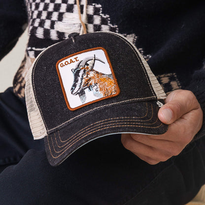 A person holds The Farm's black and beige mesh "The GOAT" hat, featuring a goat patch with "G.O.A.T." text—a standout accessory for showing off your Greatest Of All Time style.