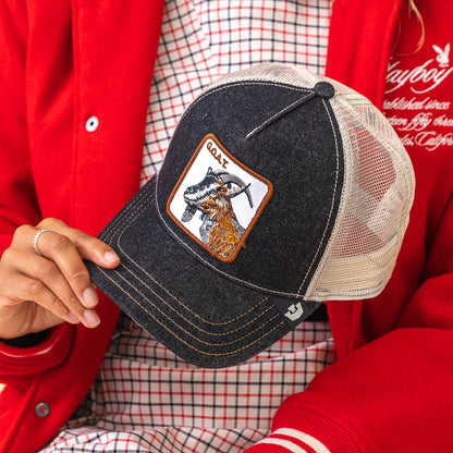 A person in a red jacket holds The Farm's best hat—The GOAT, a black and beige mesh baseball cap with a goat patch and "GOAT" embroidered on it.