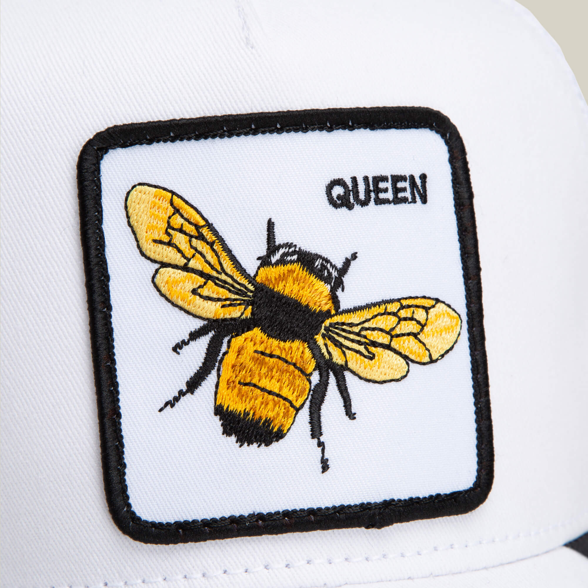 A close-up of The Farm’s “The Queen Bee” hat—a white cap featuring a patch with an embroidered yellow and black bee, a small crown above it, and the word “QUEEN” in bold black letters.