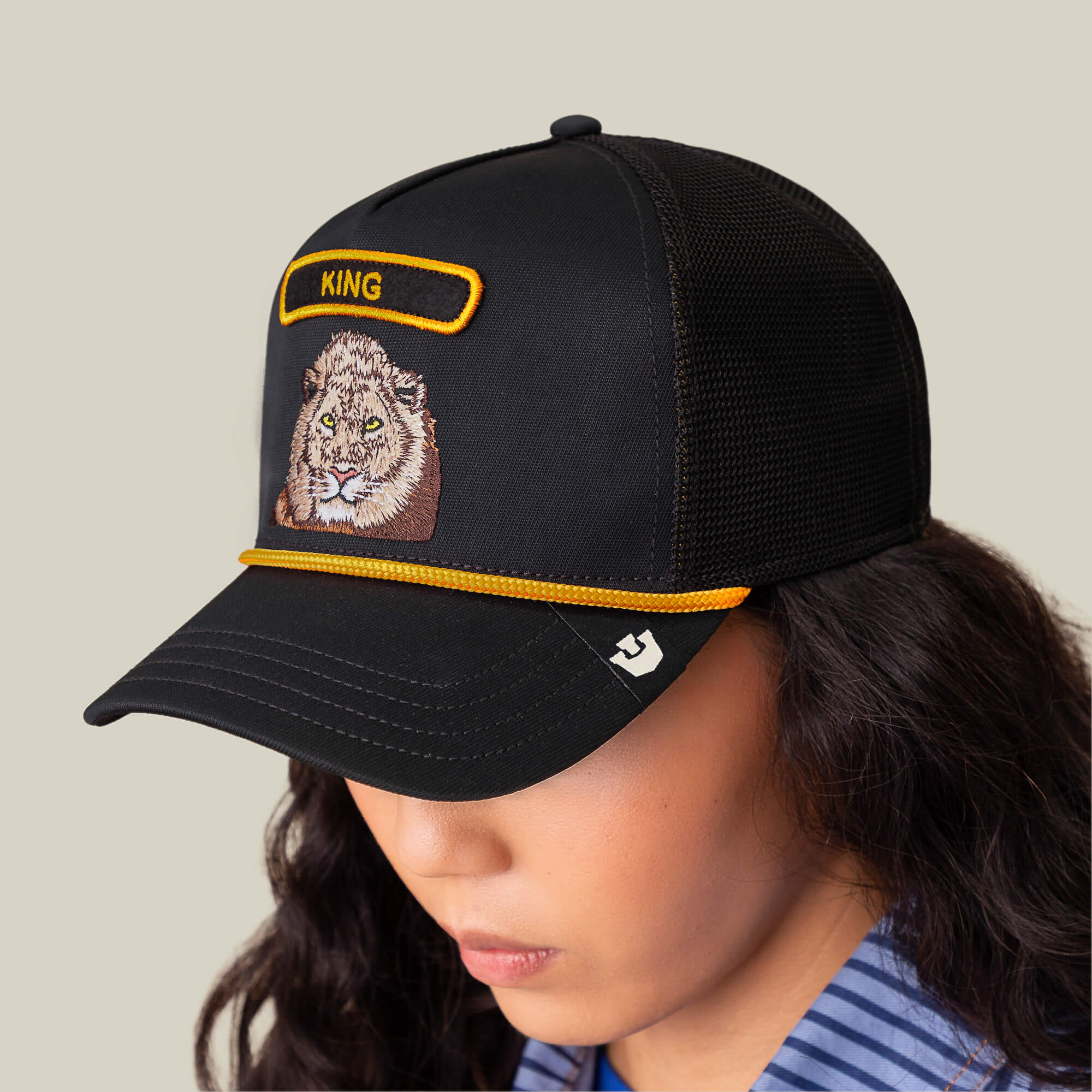 A person in The Farm’s GB2 Lion cap—black mesh design with a lion patch and KING embroidered above—looks down against a plain backdrop, symbolizing strength and greatness.