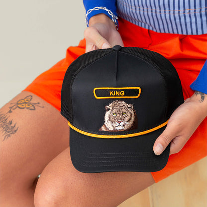 A person holds The Farm’s GB2 Lion black trucker hat, featuring an embroidered lion face and "KING" on the front. They have a butterfly tattoo on their thigh.