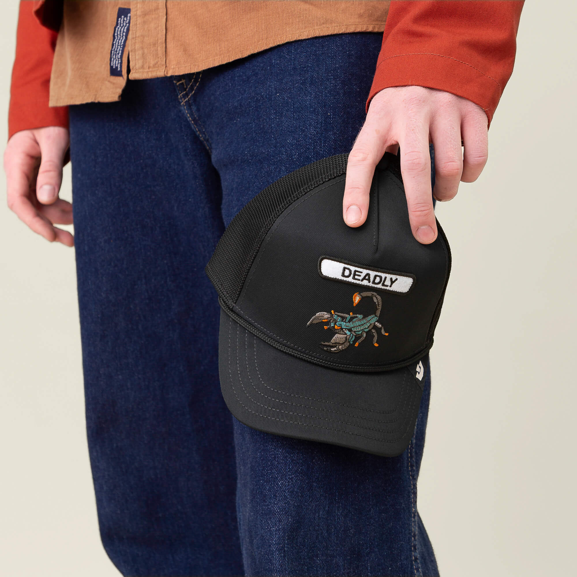 A person in jeans and a brown shirt holds the Goorin Bros. GB2 Deadly Scorp trucker hat, featuring a matte black crown, mesh back, and a scorpion patch labeled DEADLY.