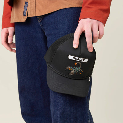 A person in jeans and a brown shirt holds the Goorin Bros. GB2 Deadly Scorp trucker hat, featuring a matte black crown, mesh back, and a scorpion patch labeled DEADLY.