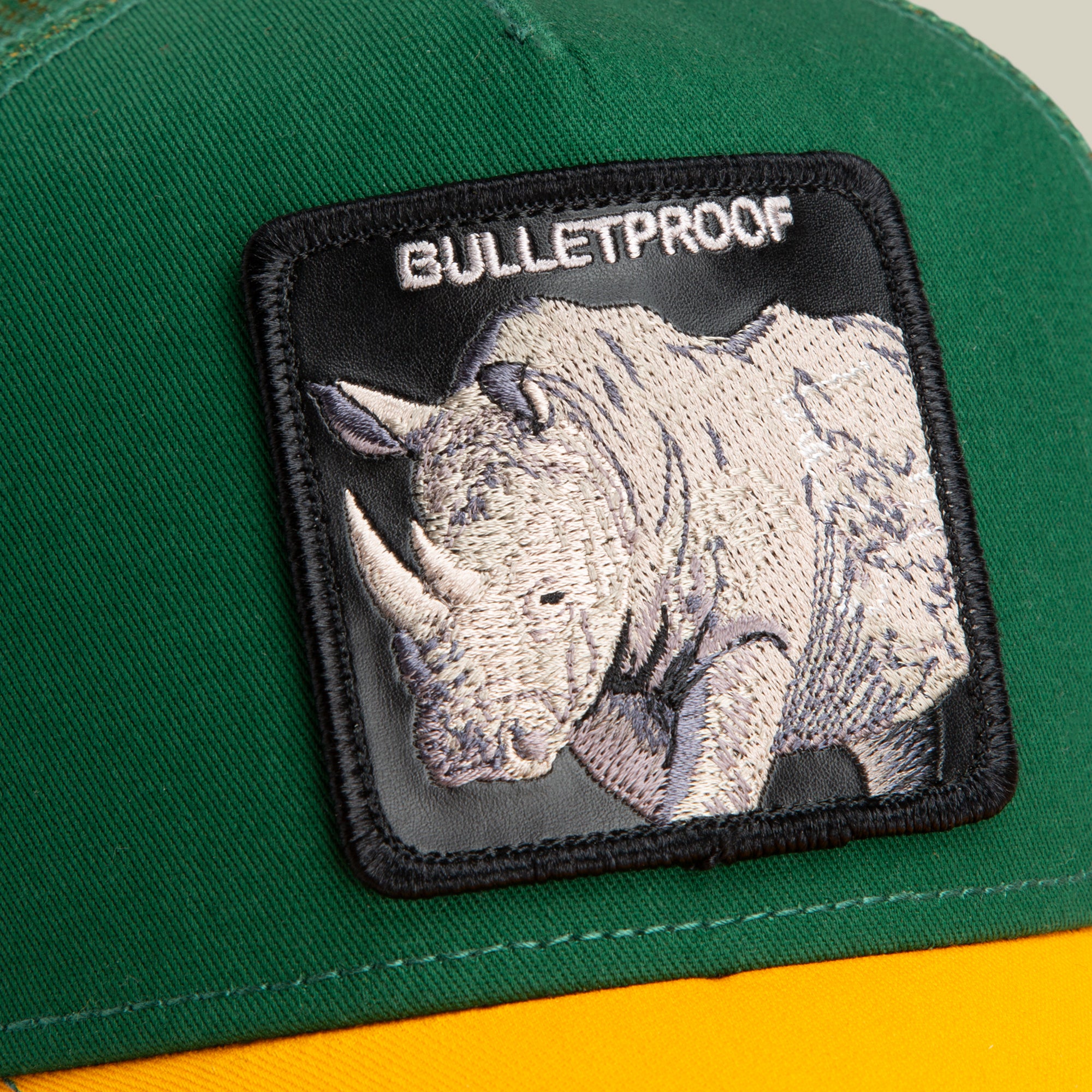 Close-up of The Farm's "The Bulletproof Rhino" cap in green and yellow, featuring a stitched rhinoceros patch and BULLETPROOF text. Please note: All sales are final.